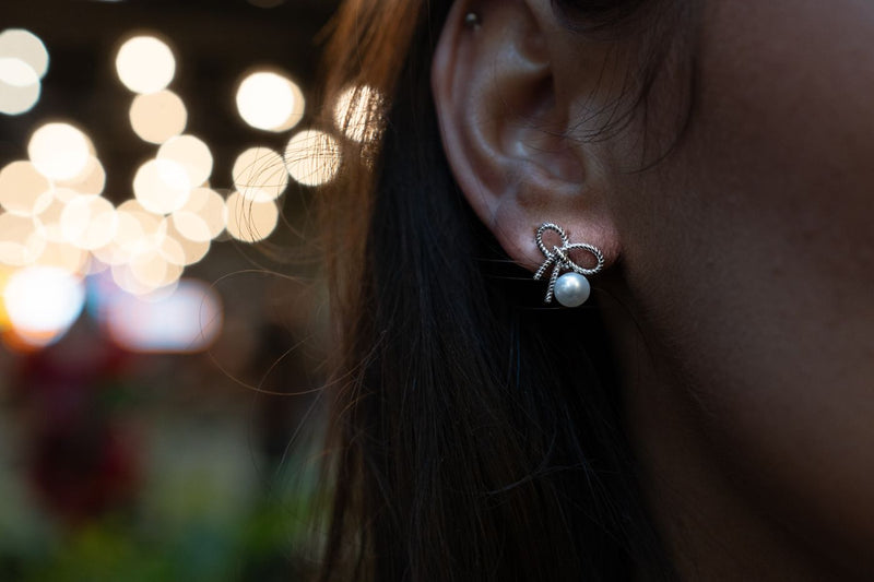 Close-up of an ear wearing a silver bow shaped earring with a pearl detail, blurred lights in the background