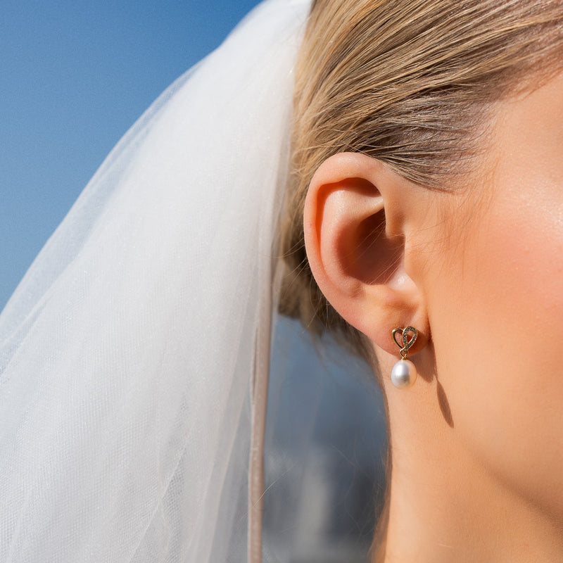 Close-up of a person wearing a pearl earring with a heart-shaped clasp, against a blurred outdoor background.
