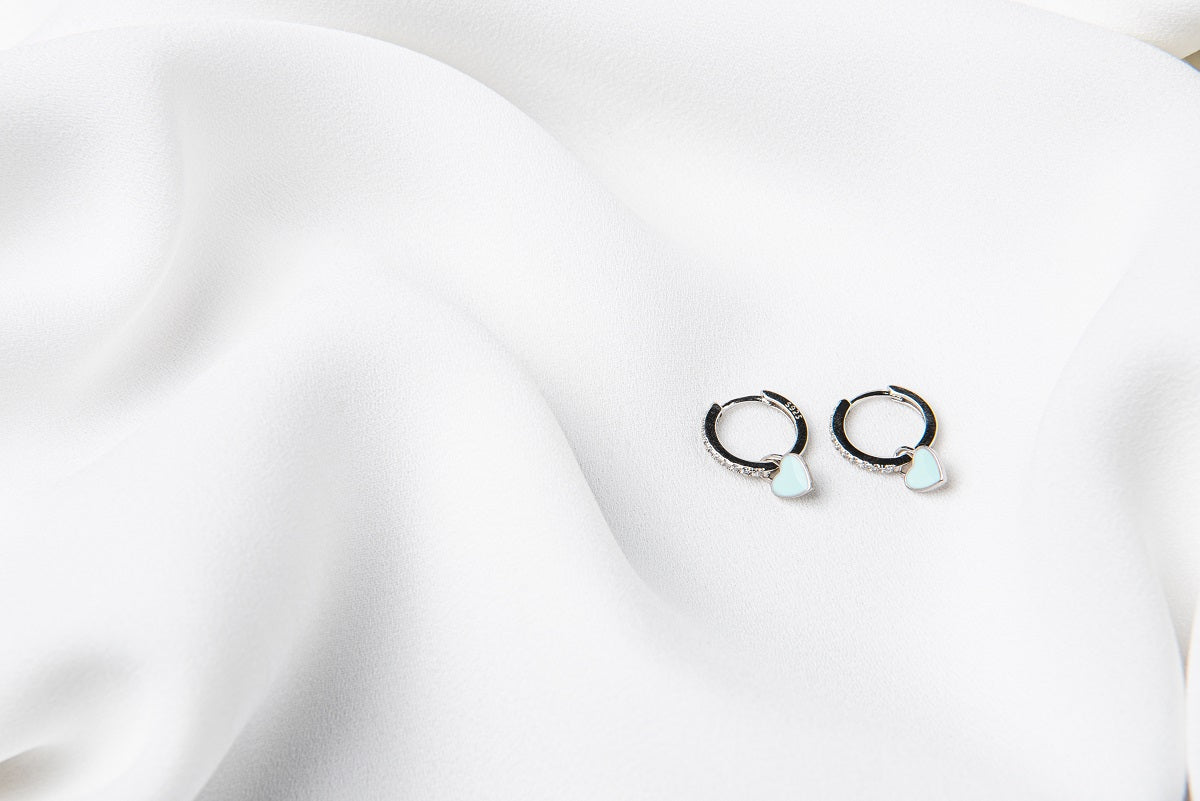 A pair of huggies earrings with blue heart-shaped dangles on a silky white background.
