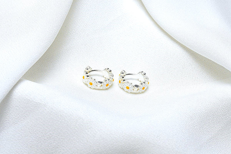 A pair of sterling silver hoop earrings with daisy-shaped embellishments, displayed on a white fabric surface.