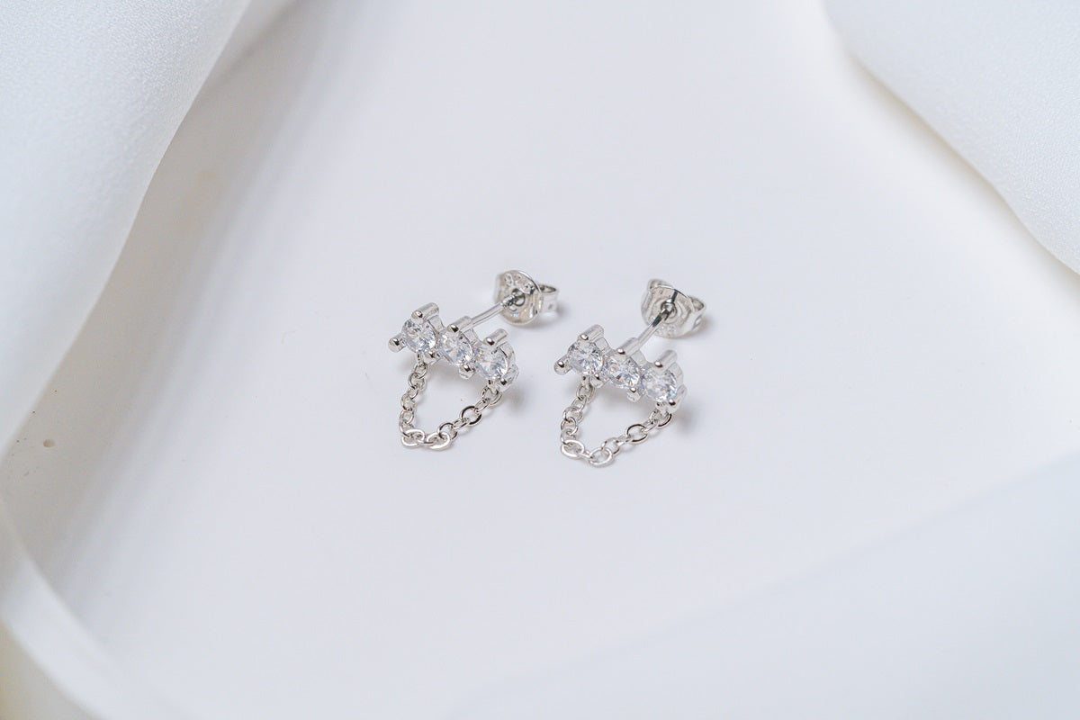 A pair of silver stud earrings with dangle loops and butterfly backs, placed on a white fabric surface.