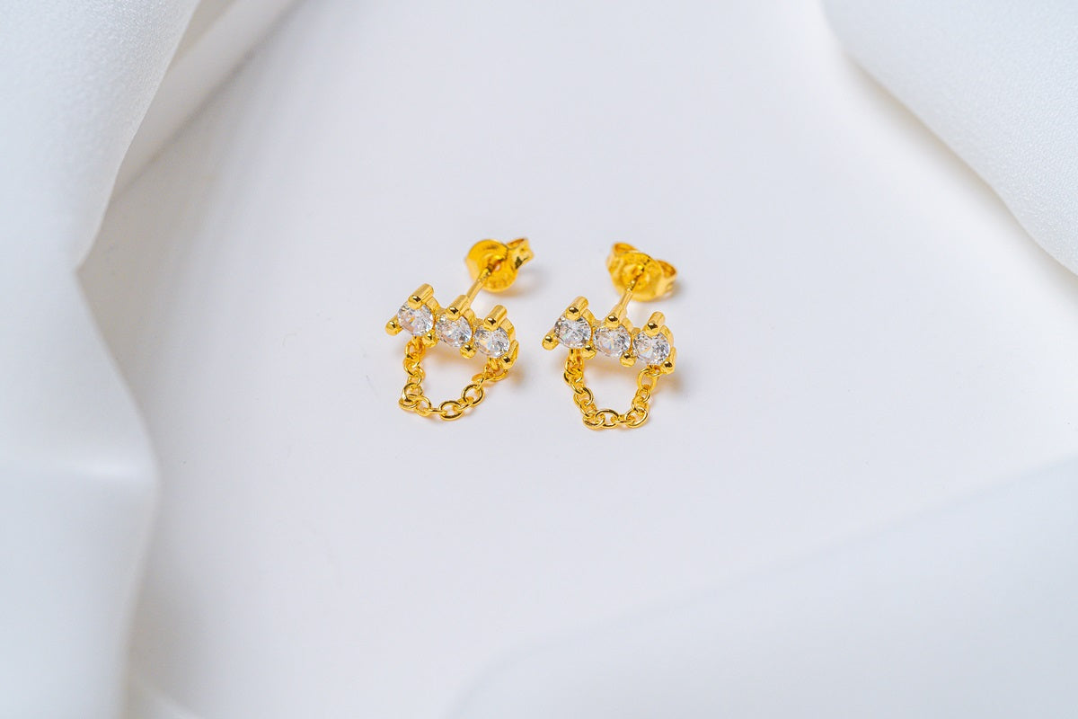 A pair of gold-colored stud earrings with dangle loops and butterfly backs, placed on a white fabric surface.
