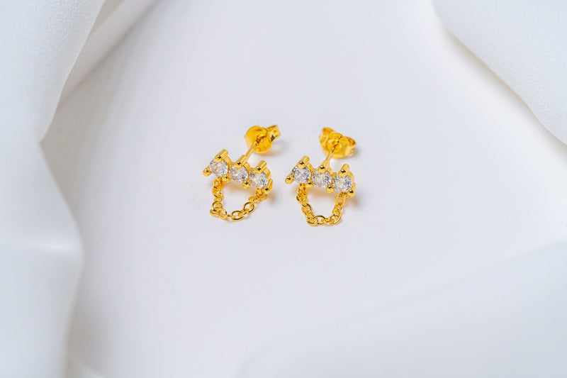 A pair of gold-colored stud earrings with dangle loops and butterfly backs, placed on a white fabric surface.