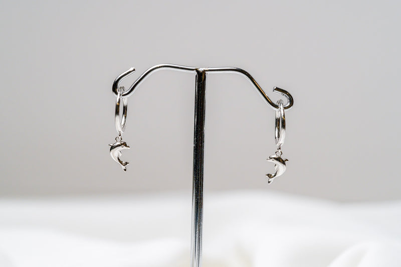 A pair of Silver hoop earrings with a dolphin design dangling from the hoops, displayed against a white background.
