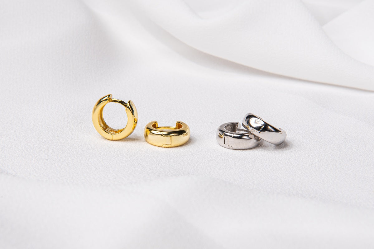Two pairs of thick huggies style earrings. One pair in sterling silver and the other pair in gold vermeil. Set on a white cloth background.