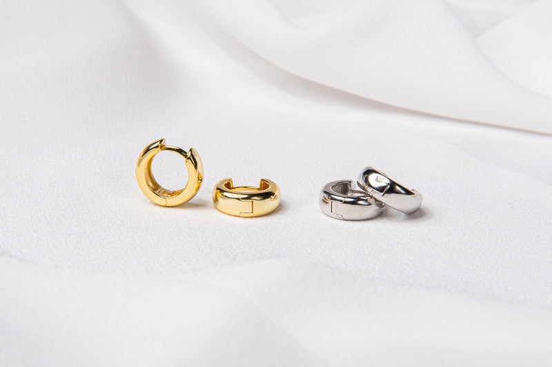 Two pairs of thick huggies style earrings. One pair in sterling silver and the other pair in gold vermeil. Set on a white cloth background.