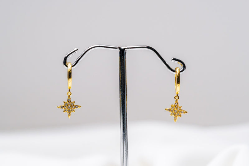 A pair of gold-colored hoop earrings with dangle diamond cubic zirconia, displayed on a silver stand against a white background.