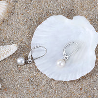 A pair of silver leverback pearl earrings on a white seashell and sandy background