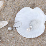 Load image into Gallery viewer, A pair of silver leverback pearl earrings on a white seashell and sandy background
