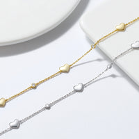 Two anklets, one plated in silver and the other in gold, each featuring a chain with three solid love heart charms.