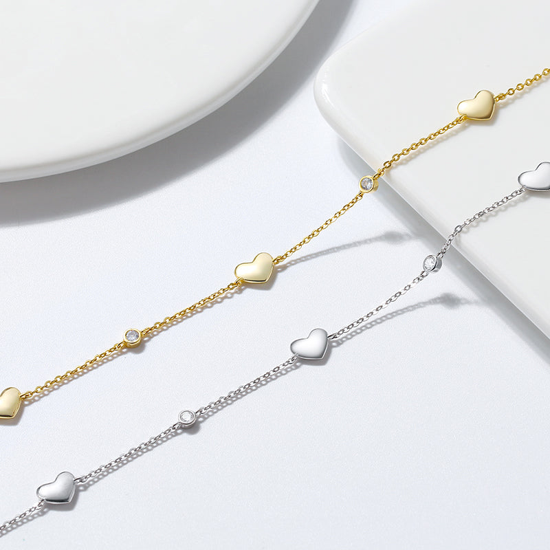 Two anklets, one plated in silver and the other in gold, each featuring a chain with three solid love heart charms.