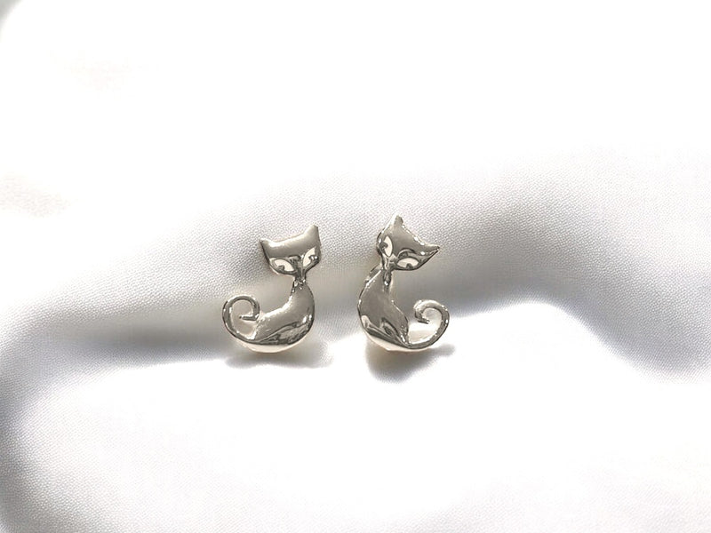 A pair of silver stud earrings featuring a cat design on the back, placed on a white fabric surface.