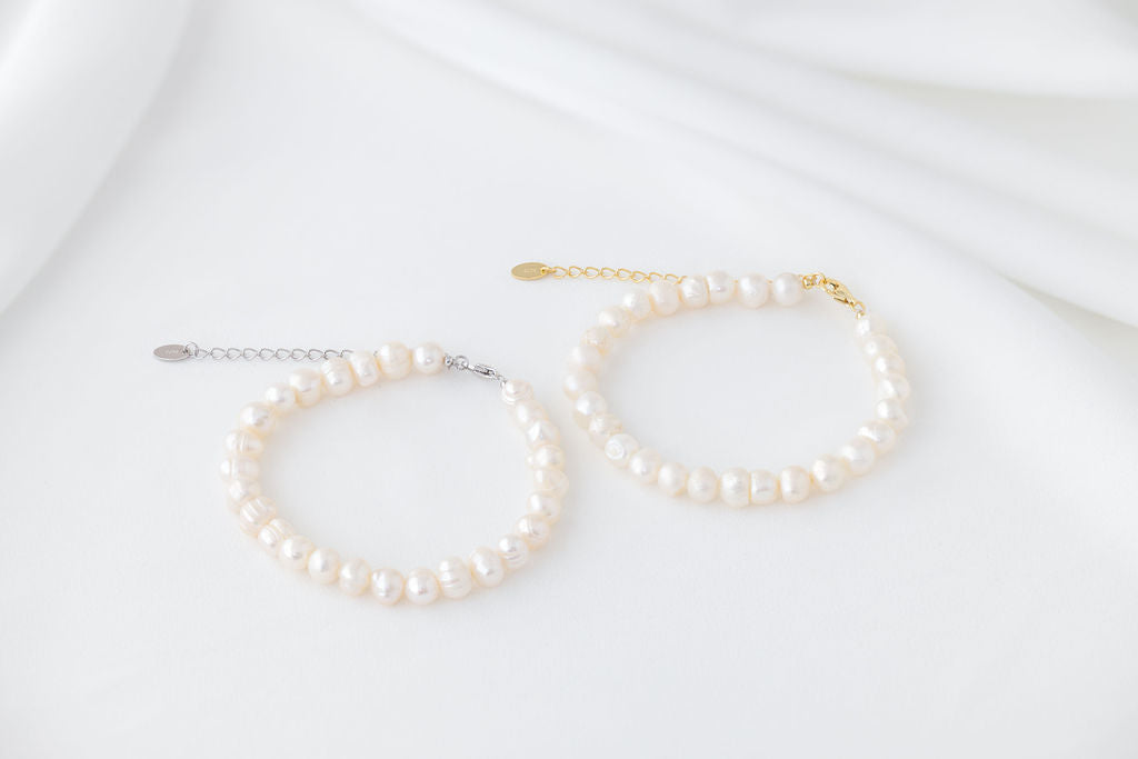 Two bracelets featureing white pearls. One with a gold chain and one with a silver chain. Displayed on a white background