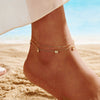 Close up of a models foot wearing a gold anklet with pendants with a beach background