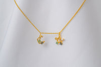 A gold-colored necklace with two blue and clear cubic zirconia pendants shaped as moons and stars, displayed on a white fabric background.