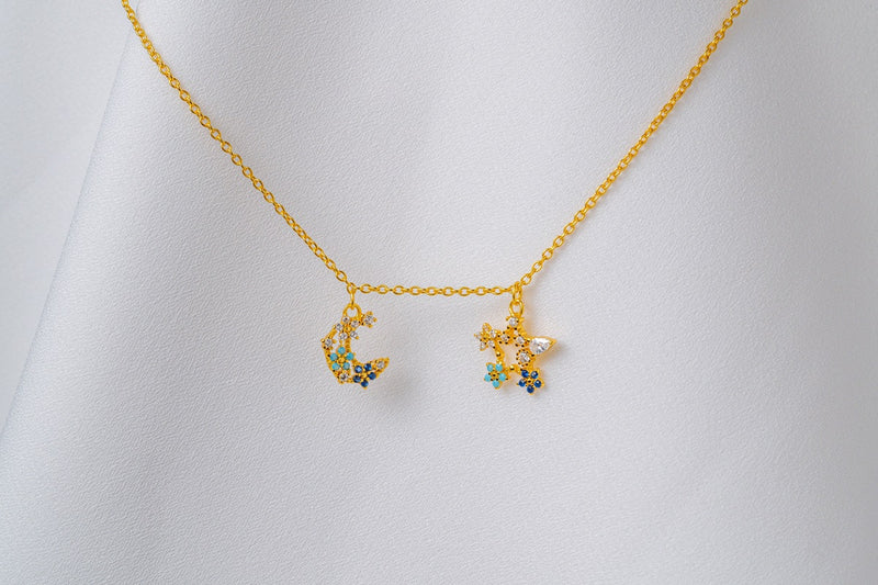 A gold-colored necklace with two blue and clear cubic zirconia pendants shaped as moons and stars, displayed on a white fabric background.