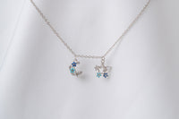 A silver necklace with two blue and clear cubic zirconia pendants shaped as moons and stars, displayed on a white fabric background.