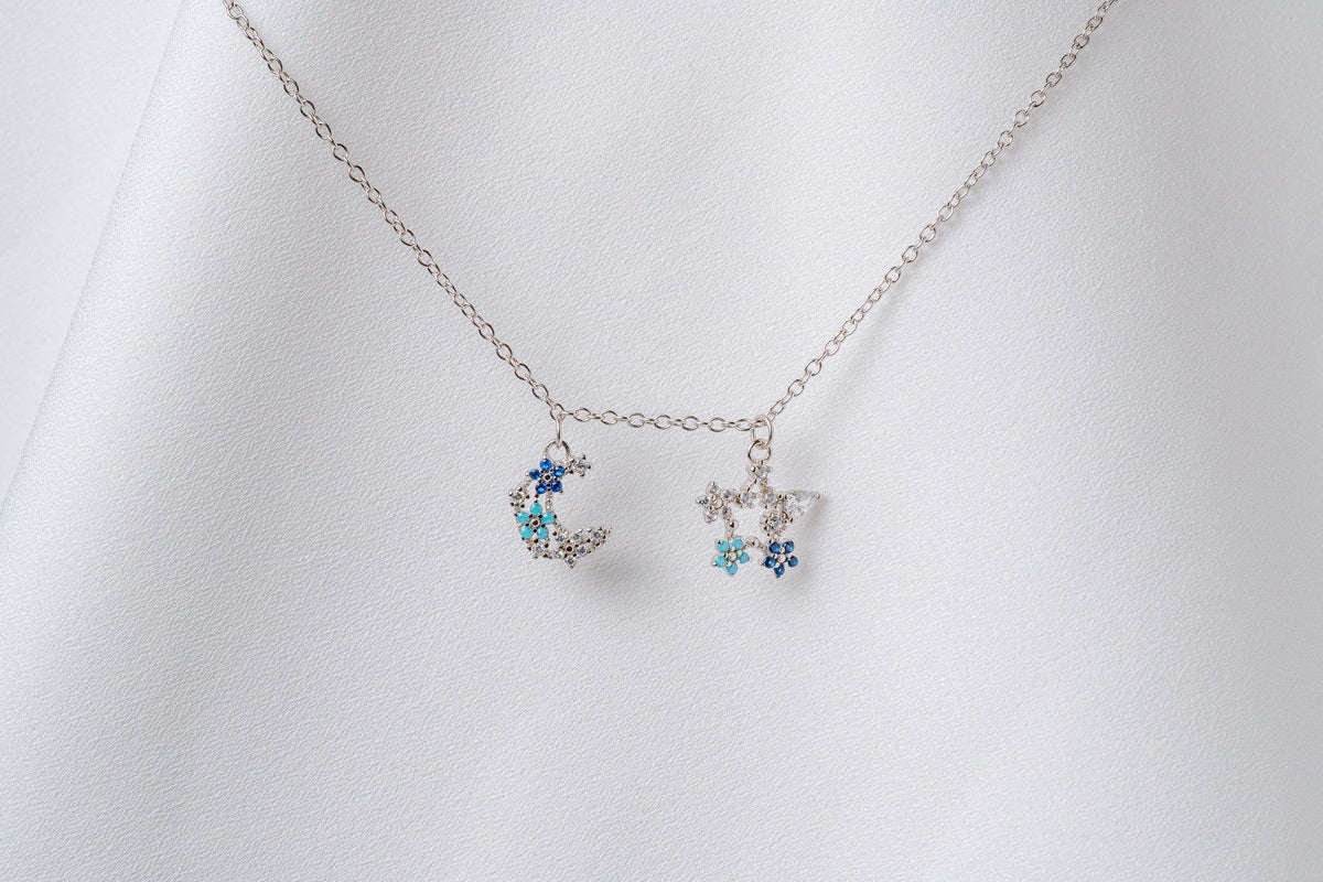 A silver necklace with two blue and clear cubic zirconia pendants shaped as moons and stars, displayed on a white fabric background.
