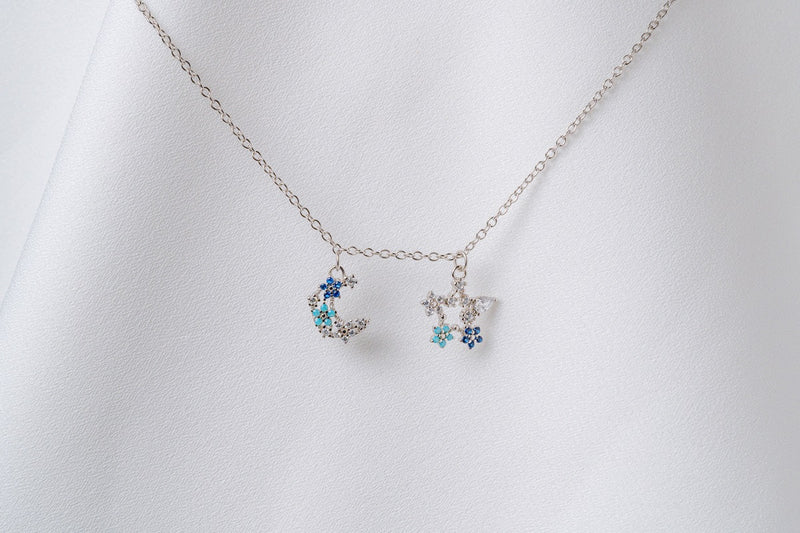 A silver necklace with two blue and clear cubic zirconia pendants shaped as moons and stars, displayed on a white fabric background.
