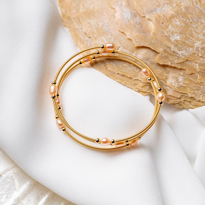 A gold wire pearl bracelet with champagne pearls, displayed on a white fabric surface next to a seashell.