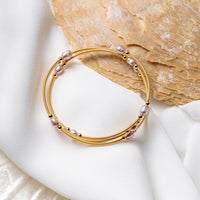 A gold wire pearl bracelet with rose pearls, displayed on a white fabric surface next to a seashell.
