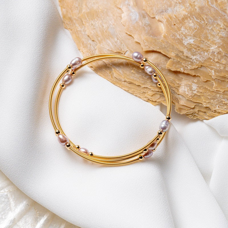 A gold wire pearl bracelet with rose pearls, displayed on a white fabric surface next to a seashell.