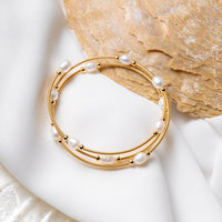 A gold wire pearl bracelet with white pearls, displayed on a white fabric surface next to a seashell.