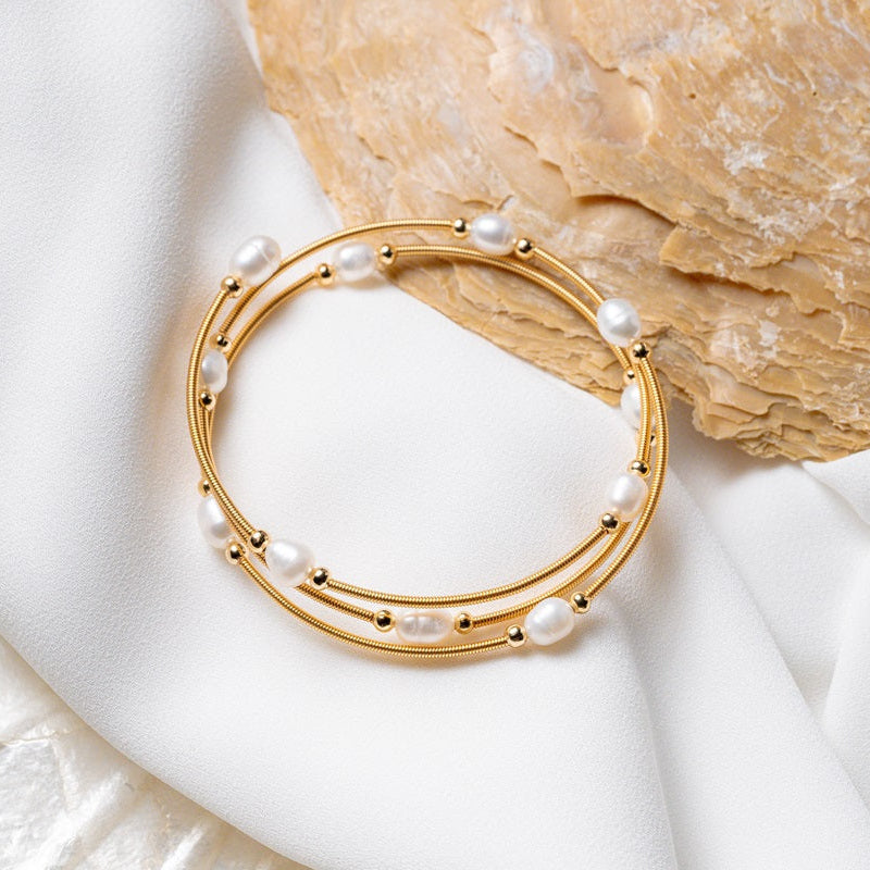 A gold wire pearl bracelet with white pearls, displayed on a white fabric surface next to a seashell.