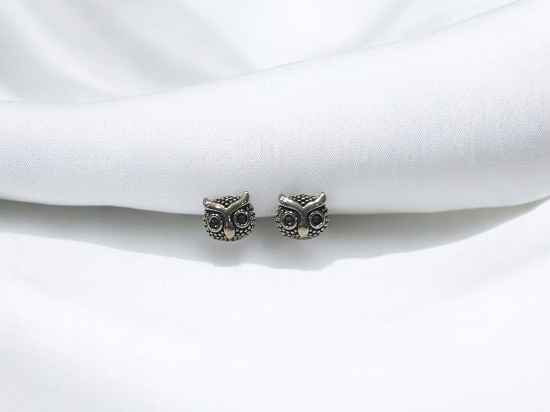 A pair of silver stud earrings with an owl design, placed on a white fabric surface.