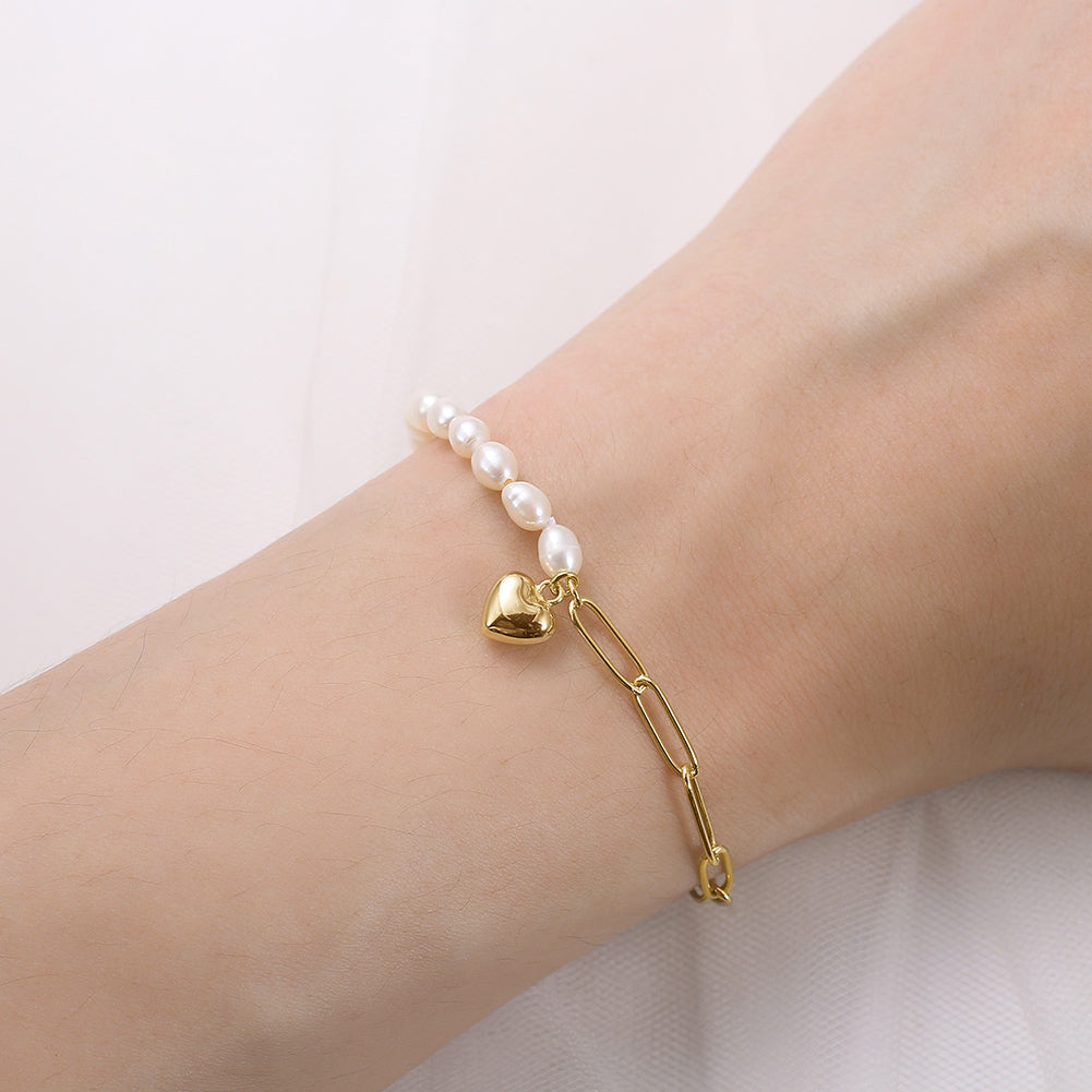Gold bracelet with heart and paperclip charm on a wrist against a light background