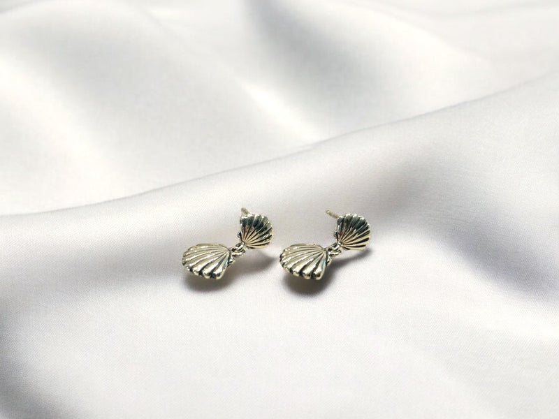 A pair of silver stud earrings with sea shell designs on a white silky surface.