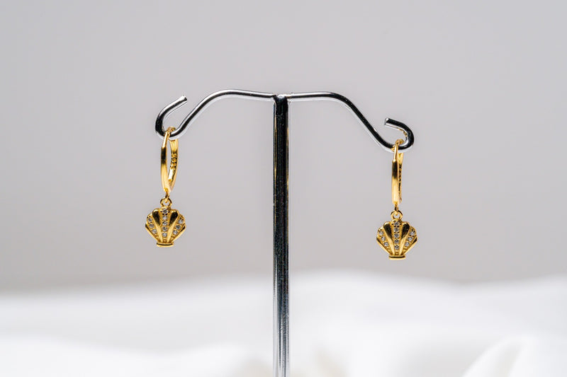 This image displays a pair of elegant shell-themed earrings, referred to as "Shell Huggies." The earrings feature a detailed and textured design that mimics the natural pattern of a shell, with a golden finish that adds a touch of sophistication.