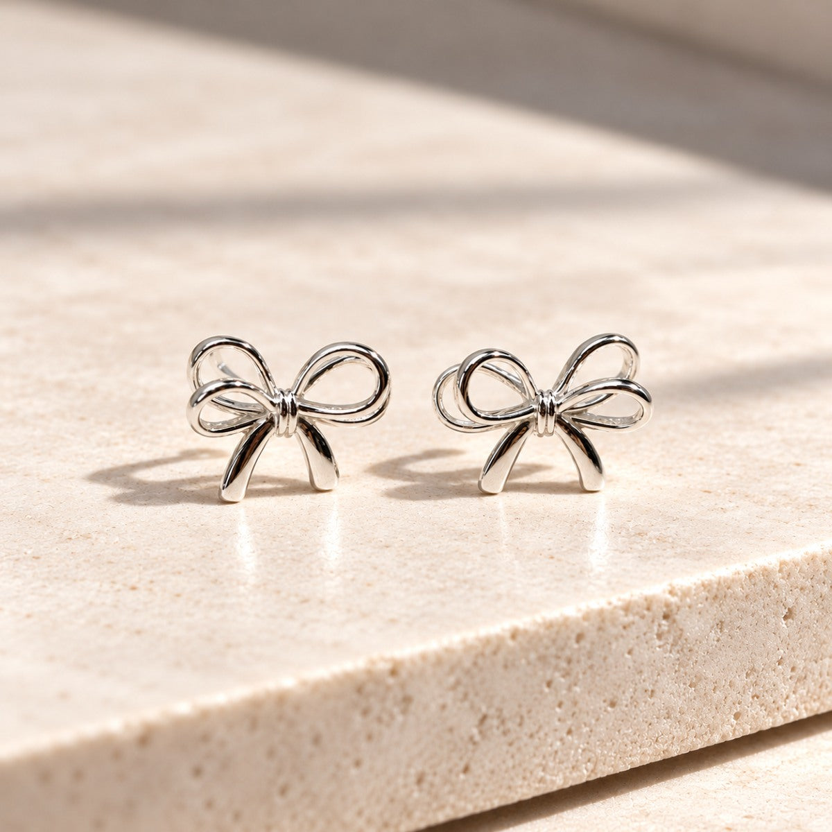A pair of sterling silver bow shaped stud earrings with butterfly backs, displayed on a white silky surface.