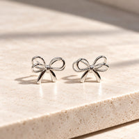 A pair of sterling silver bow shaped stud earrings with butterfly backs, displayed on a white silky surface.