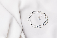 A silver bracelet with a cuban style chain design, displayed on a white fabric surface.