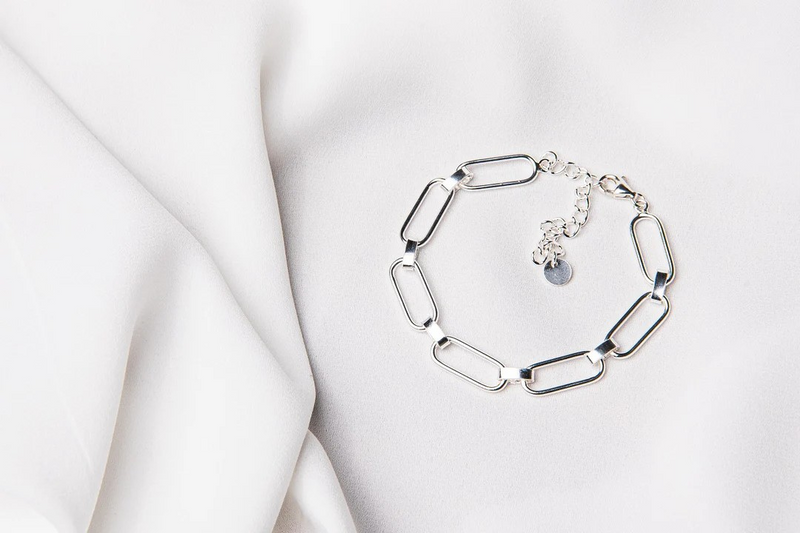 A silver bracelet with a cuban style chain design, displayed on a white fabric surface.