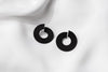 A pair of matte black earrings with a minimalist design, displayed against a white fabric background.