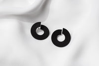 A pair of matte black earrings with a minimalist design, displayed against a white fabric background.