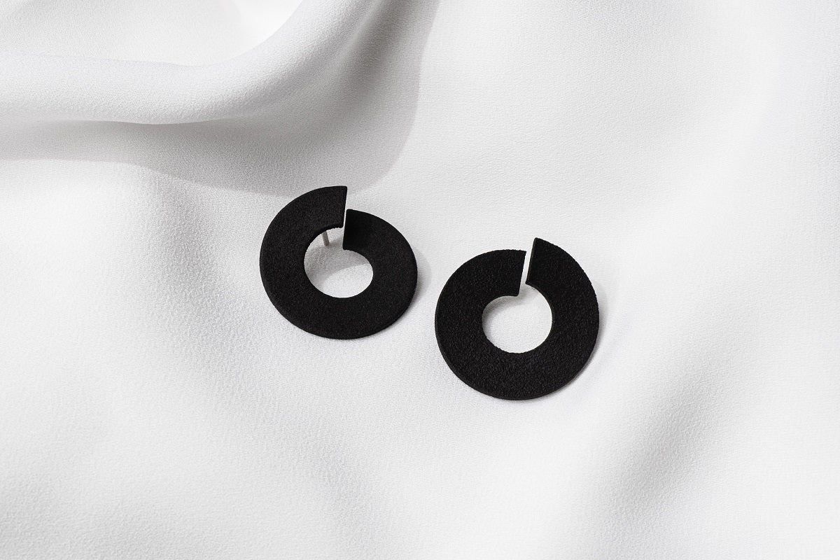 A pair of matte black earrings with a minimalist design, displayed against a white fabric background.