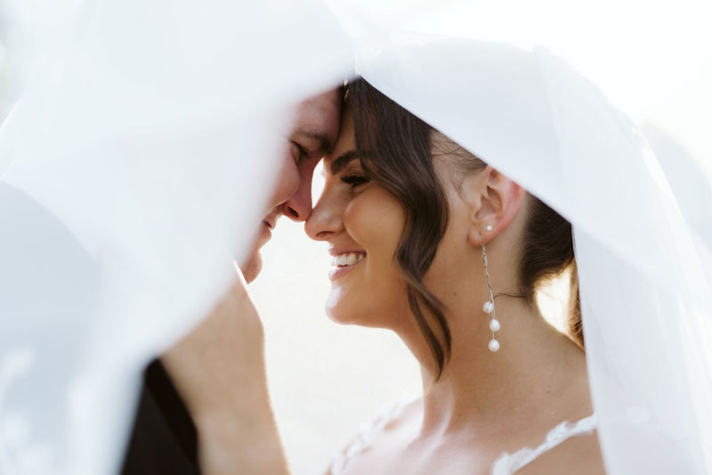 Two people standing close together with a veil draped over their heads, smiling. The bride is wearing a pair ofsilver drop earrings featuring 3 white pearls.