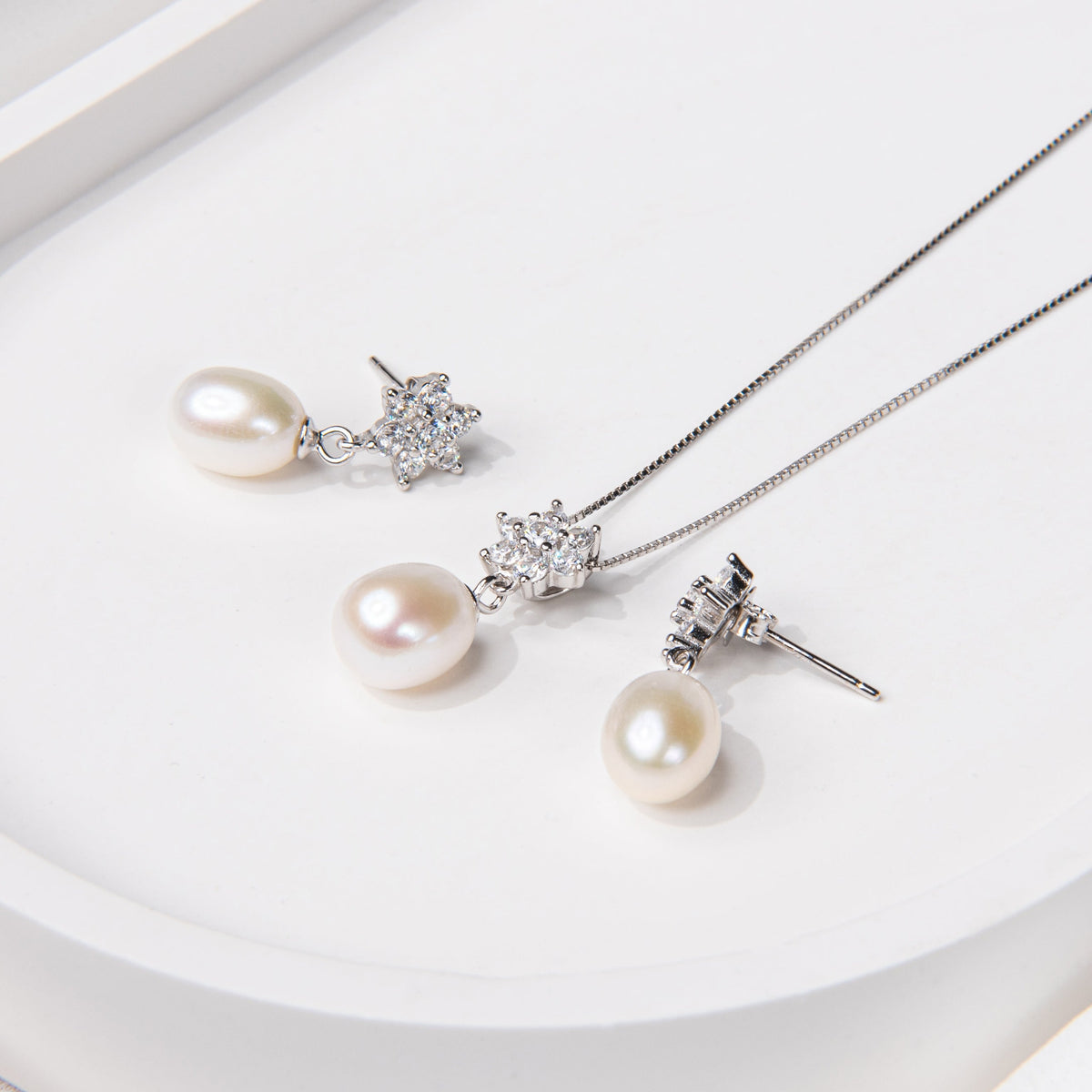 A set of sterling silver jewelry including a pendant with a drop shape pearl and flower diamond cubic zirconia, along with a pair of earrings with a similar design, displayed on a white surface.