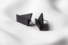 A pair of matte black, geometric drop earrings with a matte finish, lying on a white fabric surface.