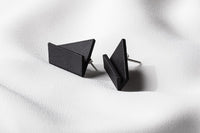 A pair of matte black, geometric drop earrings with a matte finish, lying on a white fabric surface.