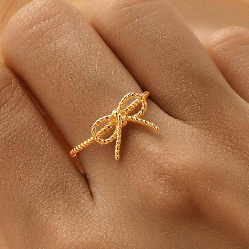 A gold vermeil ring with a bow design on a string weave band, displayed on a person's hand.