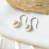 A pair of dangle pearl hook earrings in silver, placed on a white surface with a shell in the background