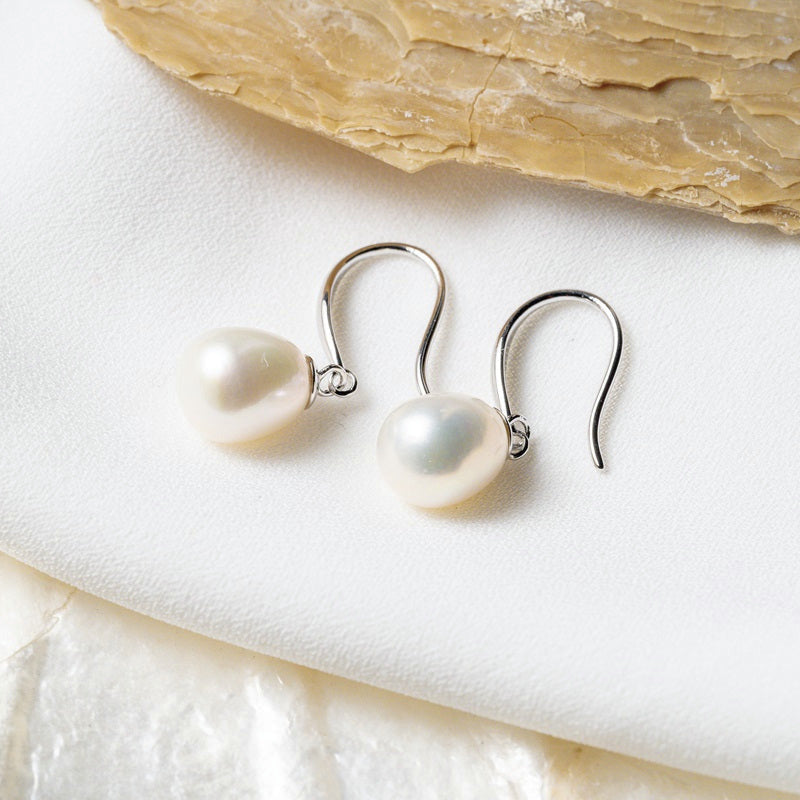 A pair of dangle pearl hook earrings in silver, placed on a white surface with a shell in the background