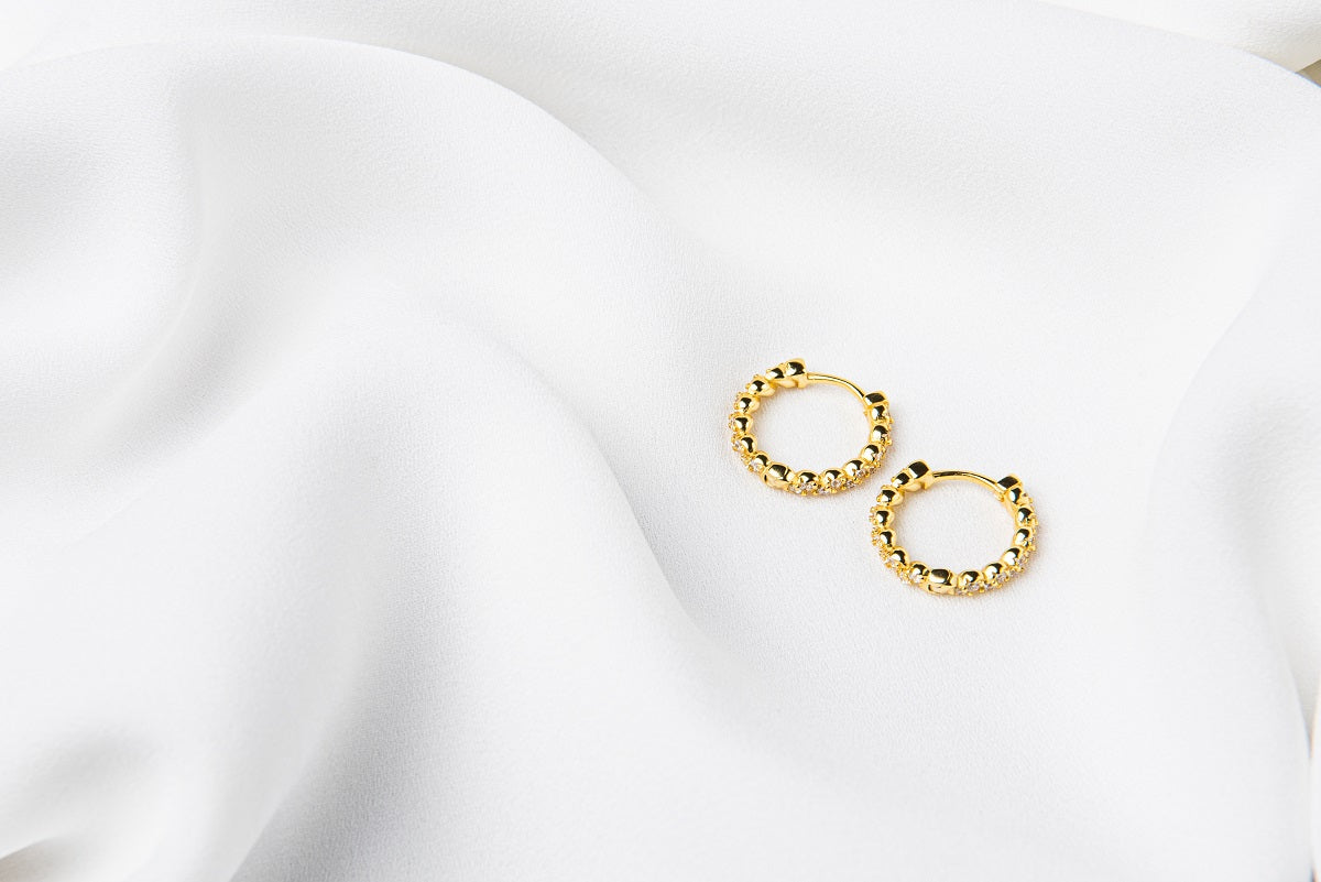 A pair of gold vermeil braided huggies earrings laid on a white silky fabric surface.