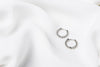 A pair of silver braided huggies earrings laid on a white silky fabric surface.