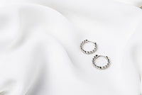 A pair of silver braided huggies earrings laid on a white silky fabric surface.