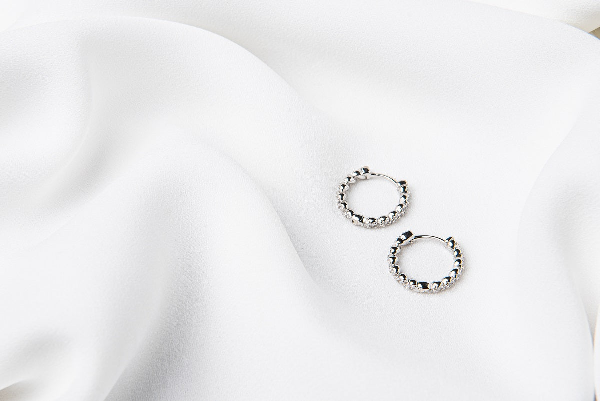 A pair of silver braided huggies earrings laid on a white silky fabric surface.